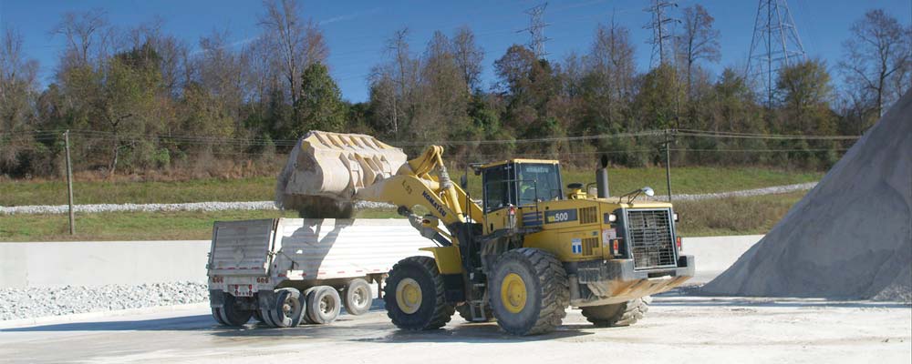 A heavy equipment operator pours ash from a front loader into the back of a semi-tractor truck